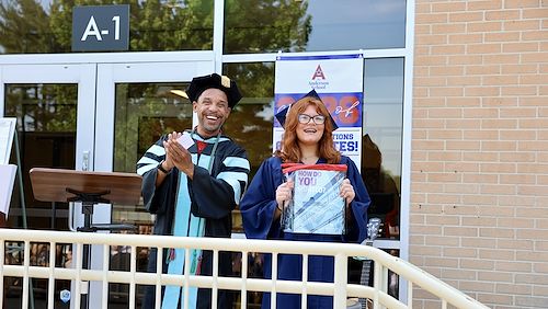 Dr. Michael Harcum dressed in graduation regalia smiles and claps his hands standing next to Anderson School graduate Kylie Eickhoff. She's holding a paper that says How Do You Montco?