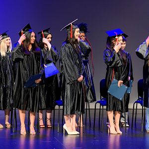 Students in black caps and gowns move the tassels on their caps during a graduation ceremony on stage.  