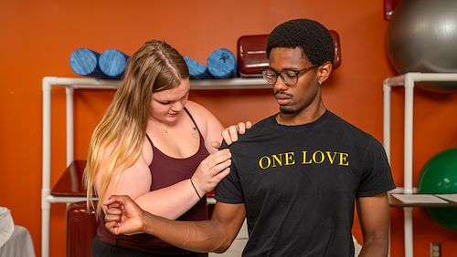 A female Physical Therapy Assistant Program student practices a technique on the shoulder of a male classmate inside a classroom. Dumbbells and exercise balls are behind them
