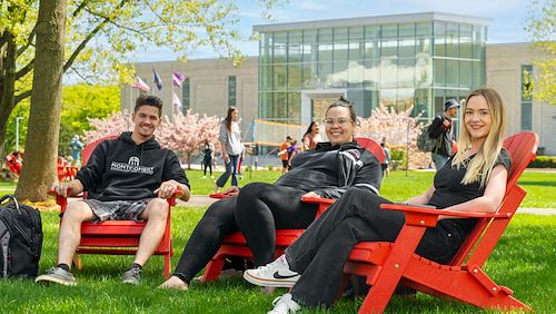 Three students sit in red chairs next to a tree. Students play volleyball behind them and College Hall is in the background.