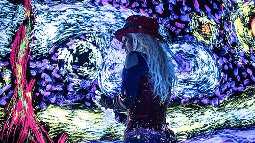 A woman wearing a top-hat paints on a black canvas in a dark room using black light paint that glows in the dark.