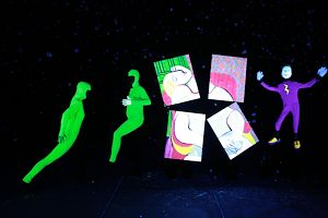 Artrageous performers wearing glow in the dark jump suits stand on stage holding Picasso’s “La Reve” in four pieces. 