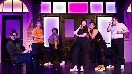 The cast of The Second City pose on stage in front of a purple lit backdrop.