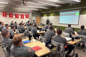 Municipal Police Academy cadets in gray uniforms are seated at desks in a classroom looking at a person giving a presentation next to a projection screen.