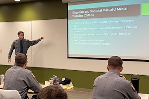 Alex Mann points to a projection screen in front of a whiteboard in a classroom. Three police cadets are seated listening to him speak.