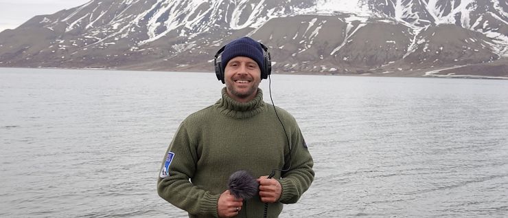 Colin Foley Colin Foley stands in front of a body of water and a snow-covered mountain holding a microphone and wearing headphones.