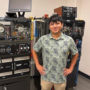 Heber Herrera stands in front of engineering equipment. His hands are on his hips.